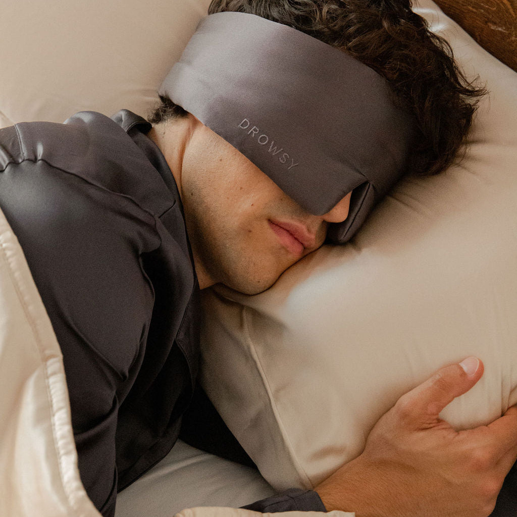A person enjoys beauty sleep with the drowsysleepco Sleep Week Ritual, wearing a gray sleep mask while resting on a white silk pillowcase and covered by a beige blanket.
