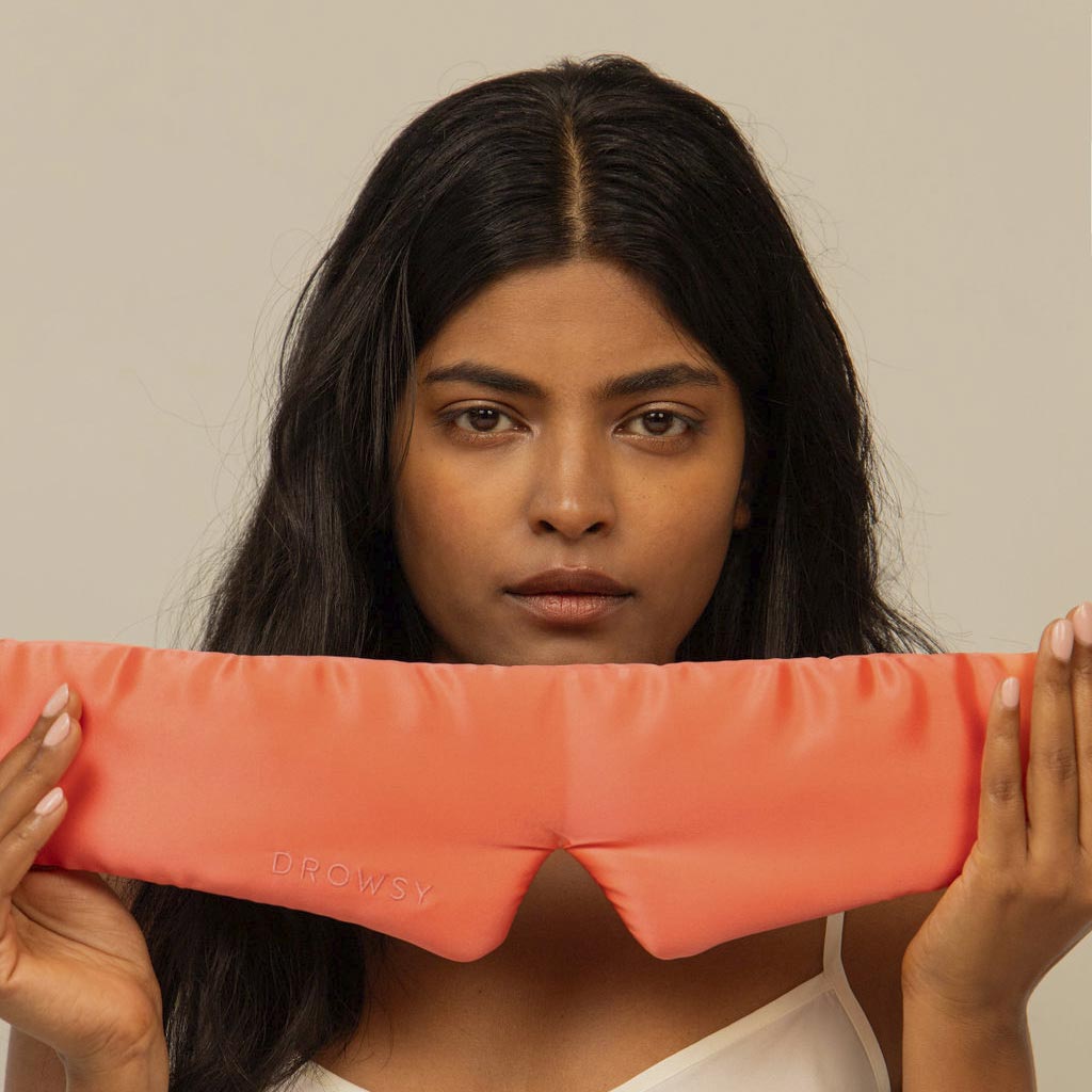 A woman with long dark hair holds a Summer Coral silk sleep mask from drowsysleepco in front of her chest, gazing at the camera with a neutral expression—ready for the ultimate beauty sleep.