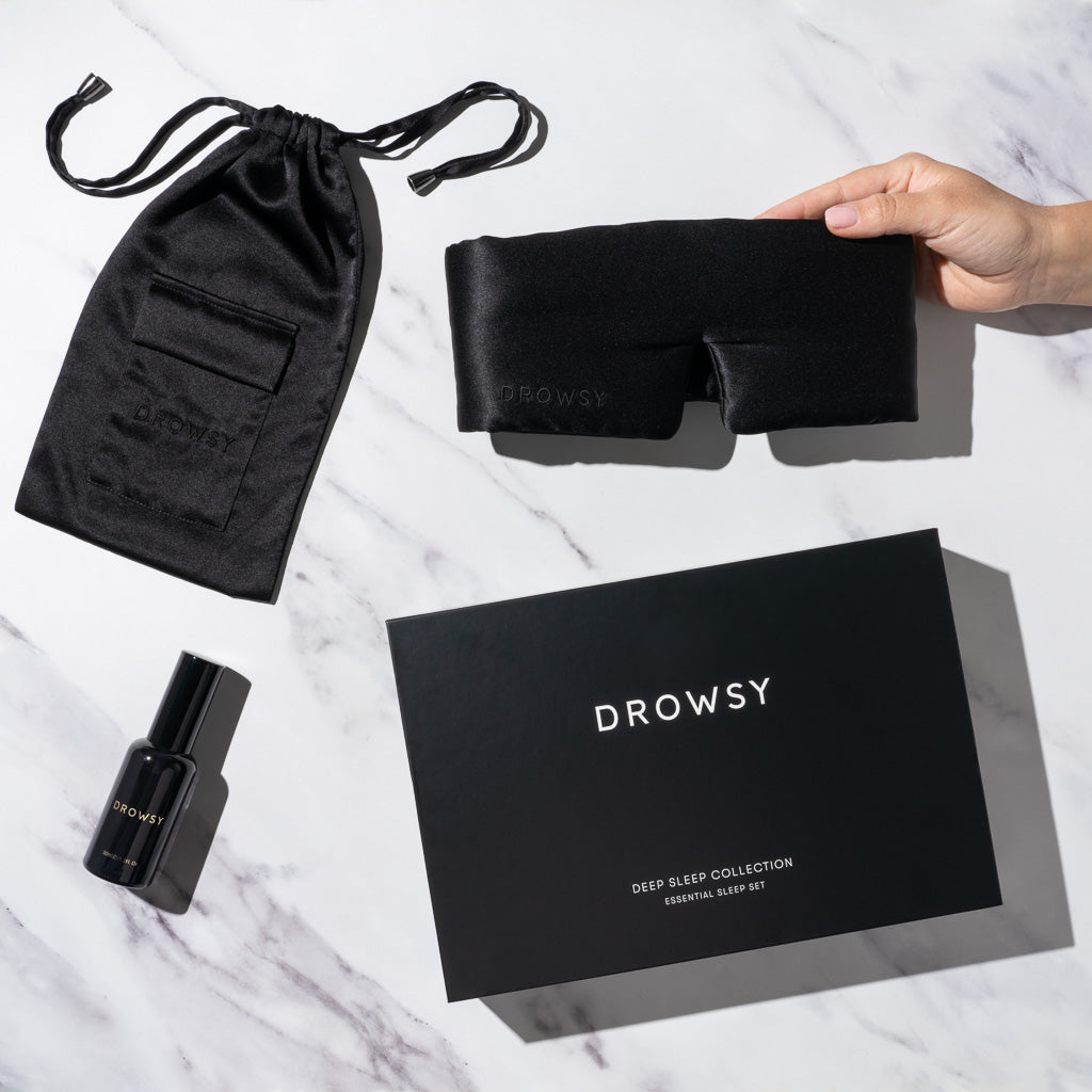 A hand holds a luxury silk sleep mask beside a black satin pouch, deep sleep pillow spray, and a black “Black Jade Deep Sleep Collection” box by drowsysleepco, all displayed on a white surface.