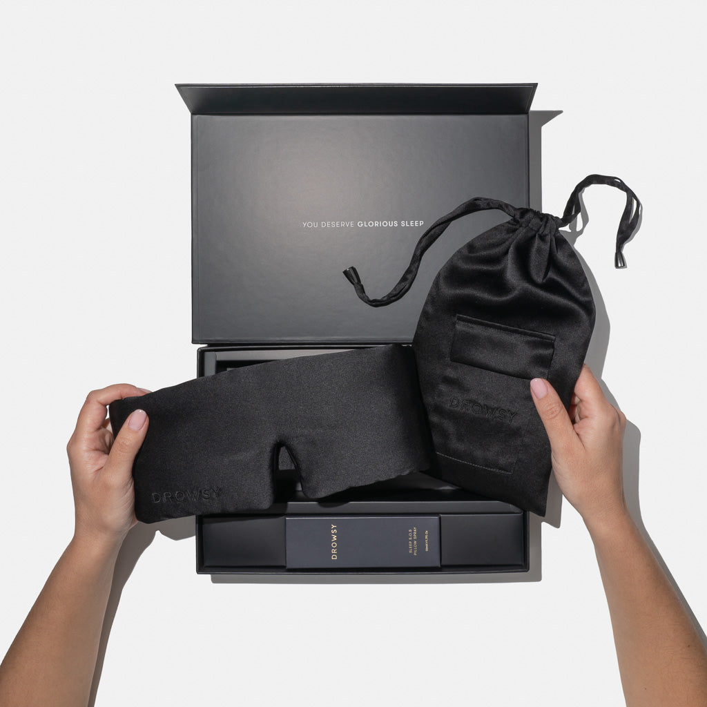 A person holds the drowsysleepco Black Jade Deep Sleep Collection silk sleep mask and matching pouch above an open black gift box with "You deserve glorious sleep" printed inside the lid.