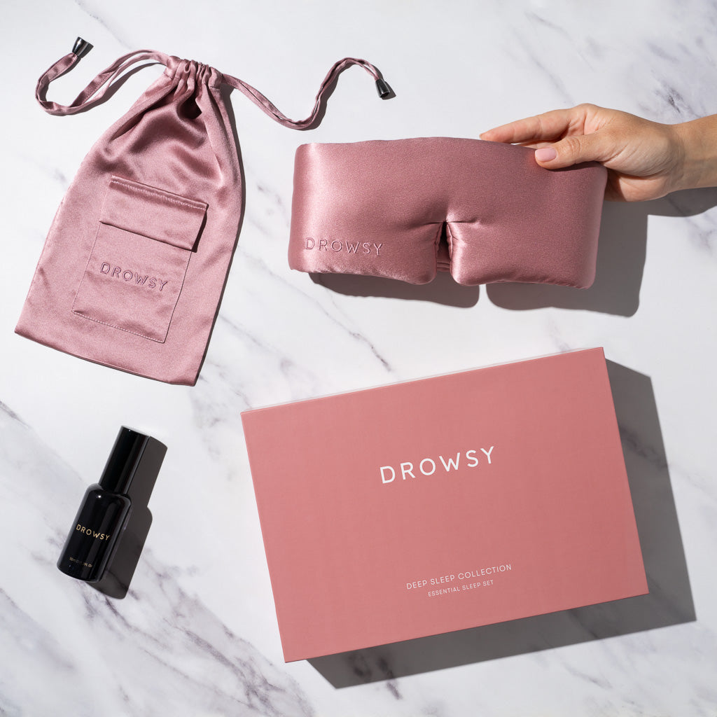 A hand holds a pink silk sleep mask by drowsysleepco beside a matching pouch, deep sleep pillow spray, and a pink box labeled Damask Rose Deep Sleep Collection on a white marble surface.