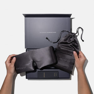 A person holds a drowsysleepco Moonlight Shadow Deep Sleep Collection silk mask and matching pouch above an open gift box reading YOU DESERVE GLORIOUS SLEEP, with a deep sleep pillow spray and other essentials inside.