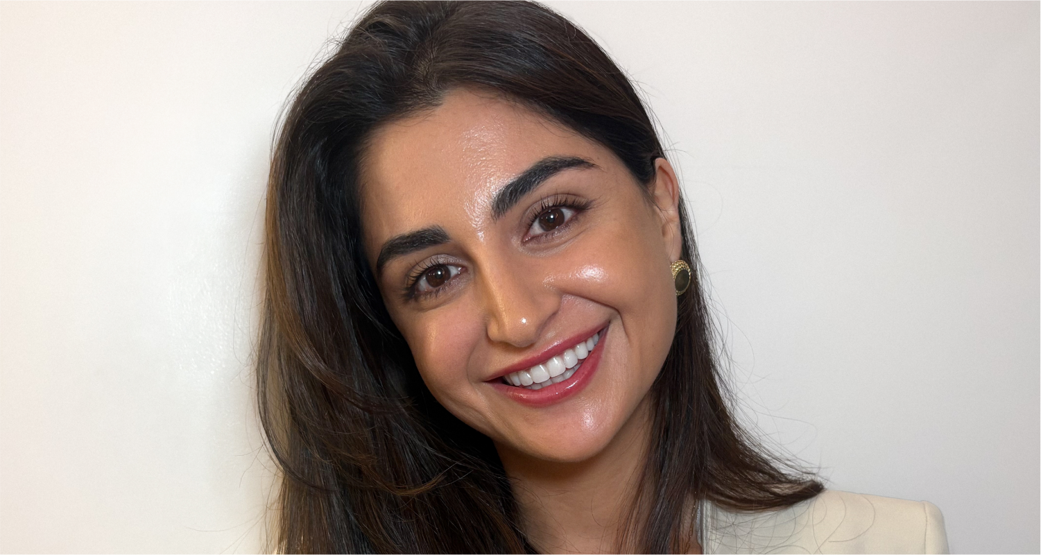 A woman with long dark hair smiles warmly at the camera, standing against a plain light-colored background. She is wearing a light-colored top and small gold earrings.