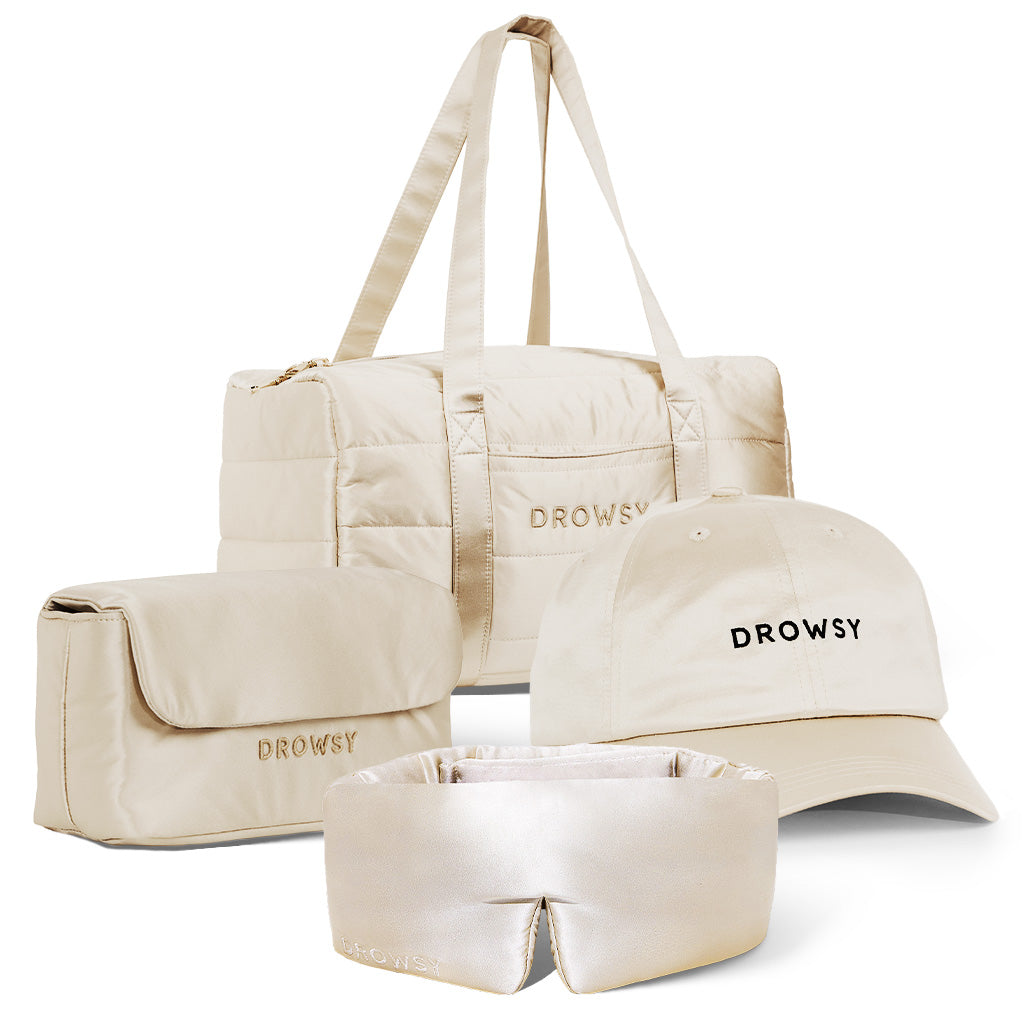 The Perfect Escape Bundle - Dusty Gold by drowsysleepco includes a beige duffel bag, toiletry pouch, DROWSY baseball cap, and a luxurious silk sleep mask—ideal for beauty sleep—all displayed on a white background.