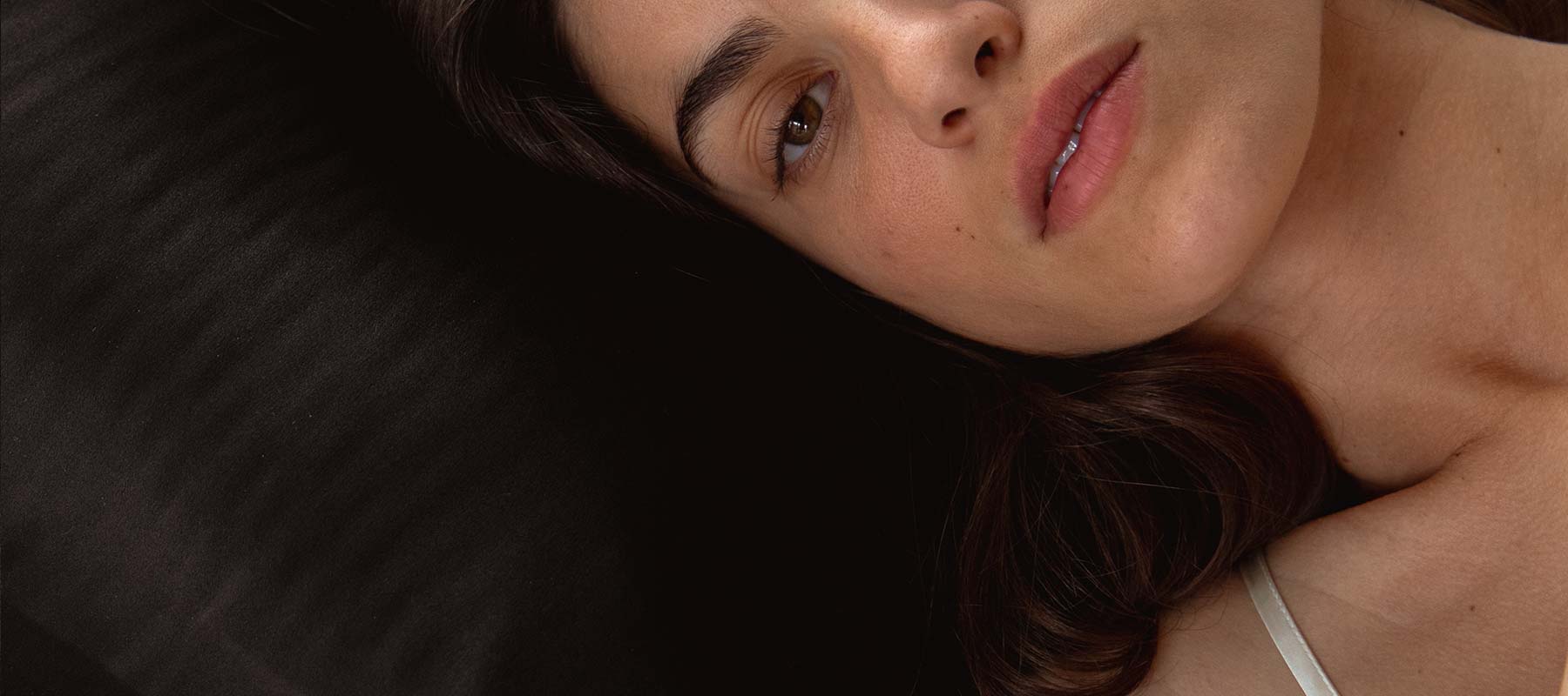 A close-up of a woman lying on her side on a black surface, looking softly at the camera. She has long dark hair and is wearing a white strap top. Only half of her face and shoulder are visible.