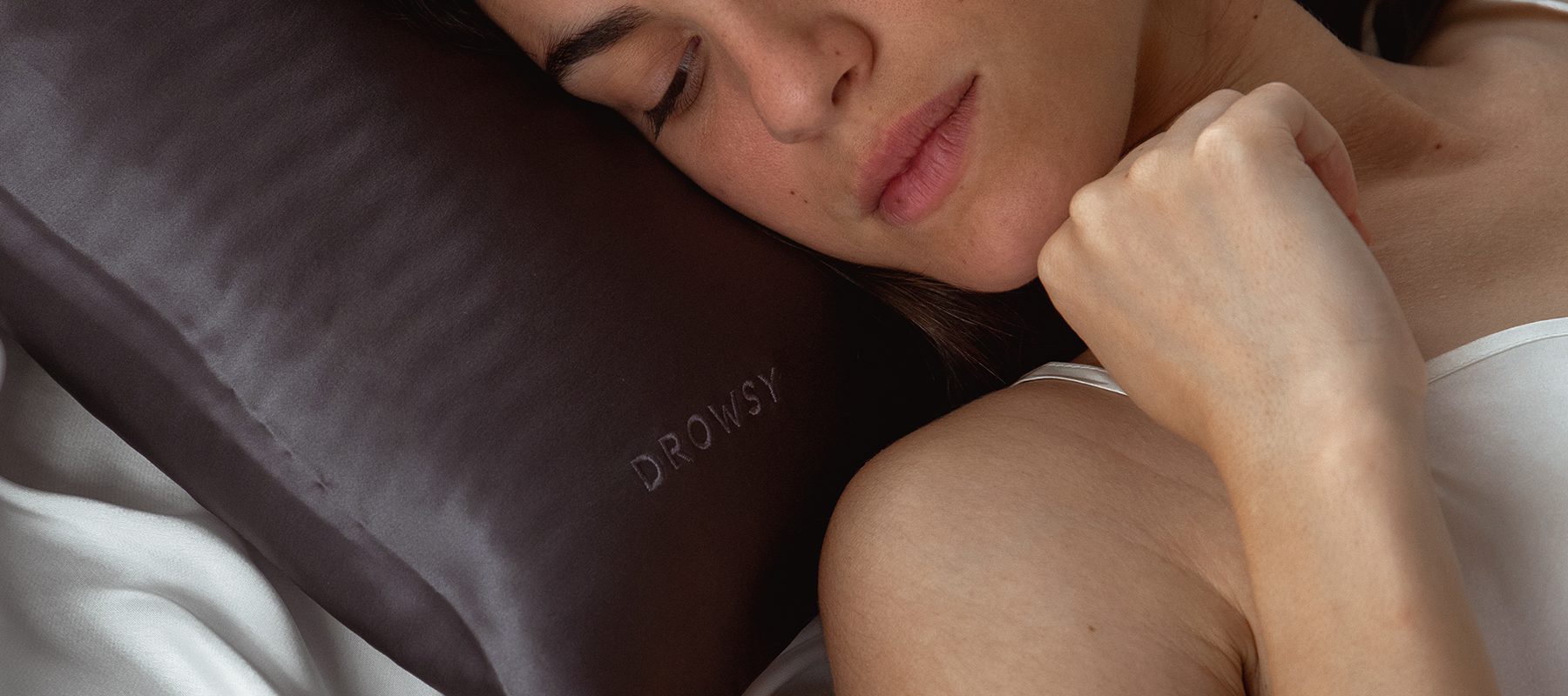 A woman in a white top lies on her side in bed, resting her head on a dark silk pillow embroidered with the word DROWSY. She appears peaceful and relaxed.