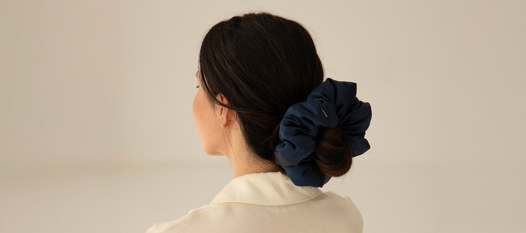 A woman with dark hair, styled in a low bun, wears a large navy blue scrunchie. She is shown from behind and is wearing a cream-colored top against a plain, light background.
