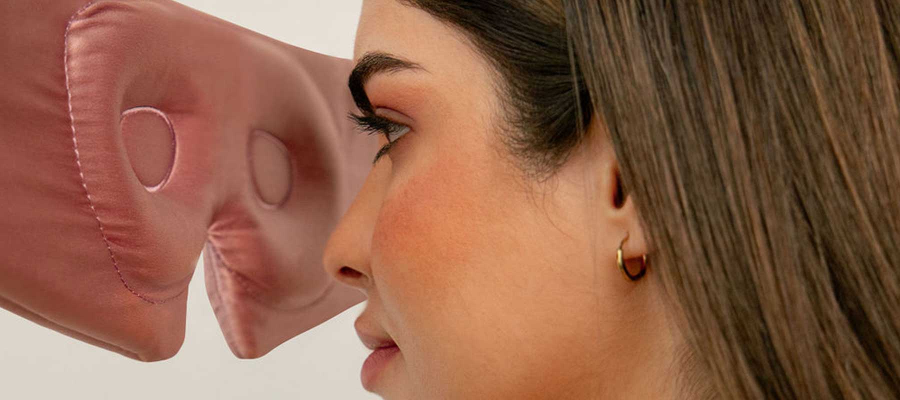 A woman with straight brown hair and gold hoop earrings faces a large, pink, artificial nose positioned close to her face.