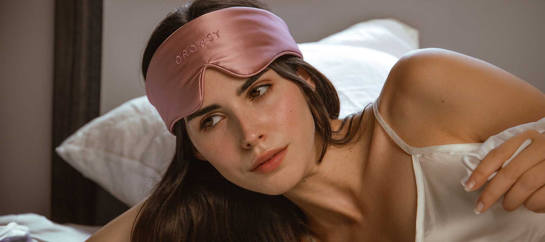 A woman in a white satin camisole lies on a bed, wearing a pink sleep mask pushed up on her forehead. She looks thoughtful and relaxed. Pillows and white bedding are visible in the background.