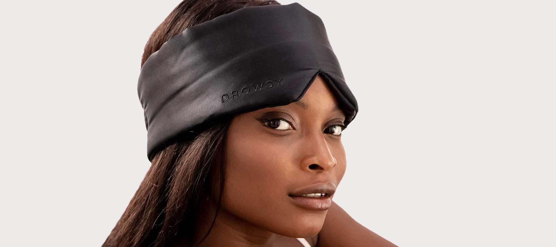 A woman with long dark hair wears a black silk sleep mask pushed up on her forehead, looking confidently at the camera against a plain light background.