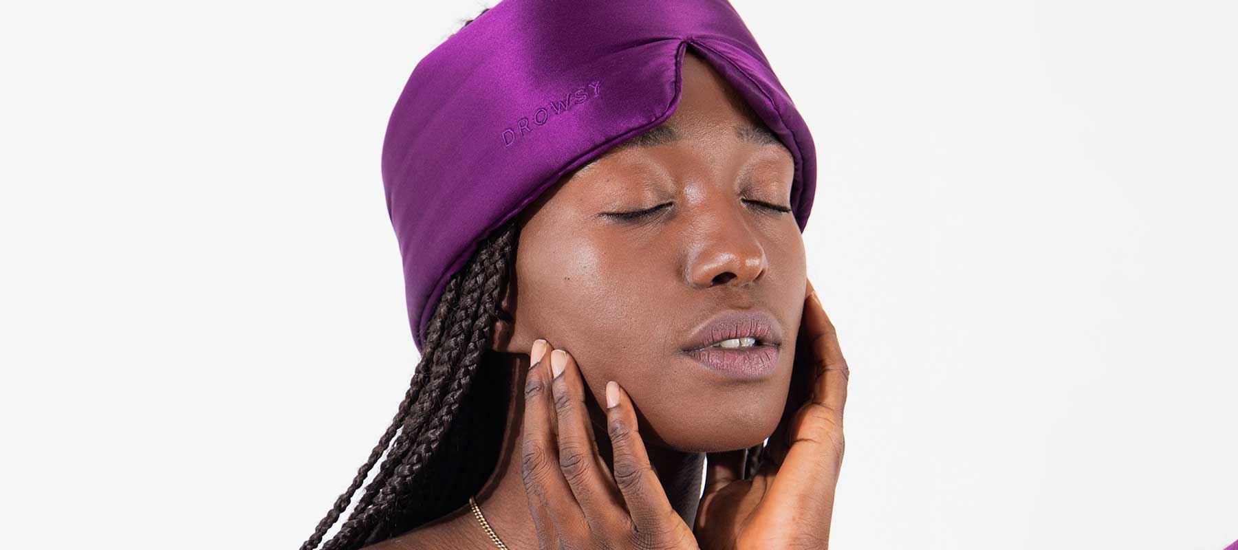 A person with braided hair wears a purple satin eye mask and gently touches their face with both hands, eyes closed, appearing relaxed against a plain white background.