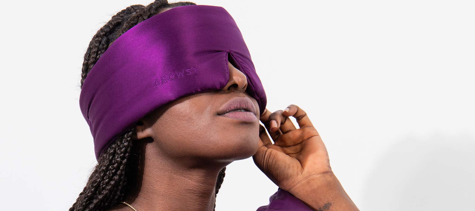 A person with braided hair wearing a large, deep purple silky sleep mask, covering their eyes and forehead, with one hand gently touching the side of their face.
