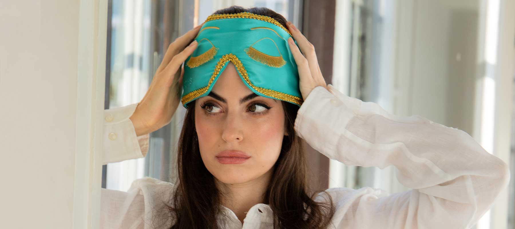 A woman with long dark hair adjusts a turquoise sleep mask with gold trim on her head while wearing a white blouse, looking off to the side.
