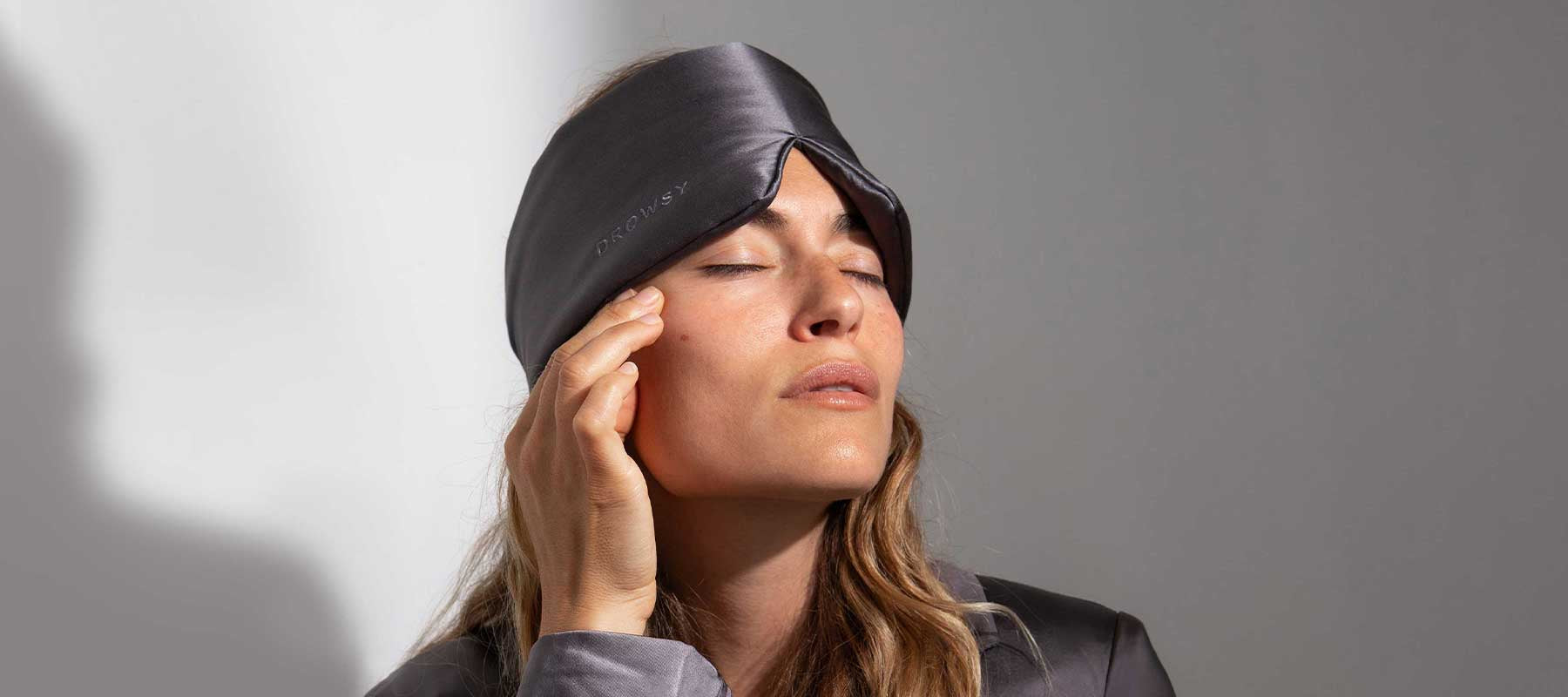 A woman wearing a dark gray sleep mask and matching pajamas lightly touches her temple with her eyes closed, appearing calm and relaxed against a plain background.