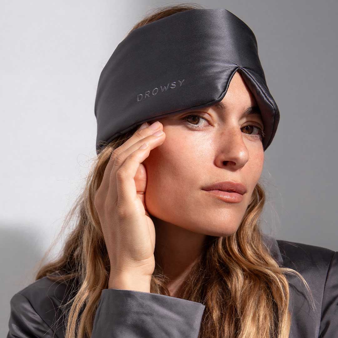 A woman with long, wavy hair wears a drowsysleepco Midnight Blue sleep mask from the 2-Pack Sleep Masks (Midnight Blue + Moonlight Shadow), pushed up on her forehead as she calmly touches her face and looks at the camera.