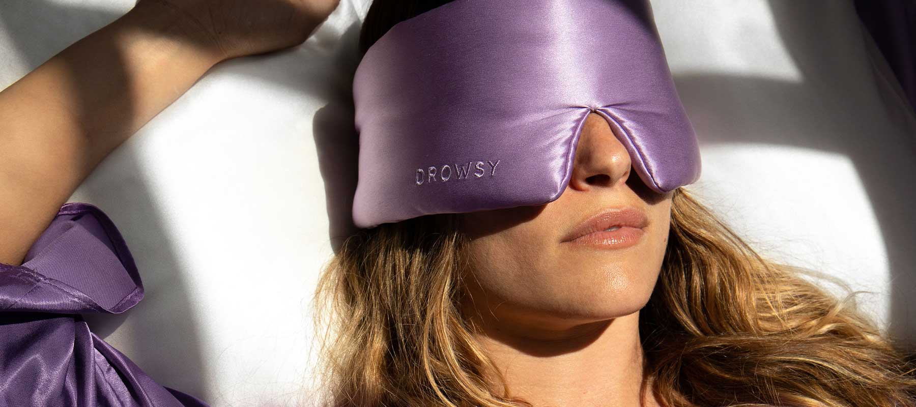 A woman wearing a shiny lavender eye mask labeled DROWSY lies on her back in bed, with sunlight casting soft shadows across her face and purple robe.