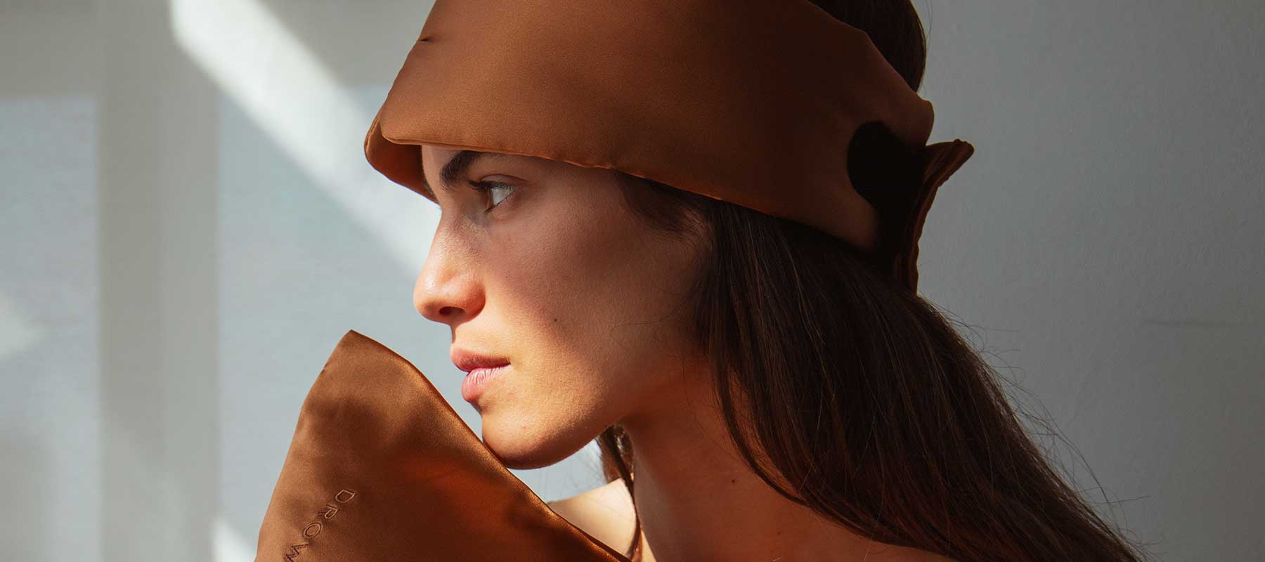 A woman with long brown hair faces sideways, wearing a brown silk eye mask on her head and holding a matching silk pillow. Sunlight casts soft shadows on her face and the wall behind her.