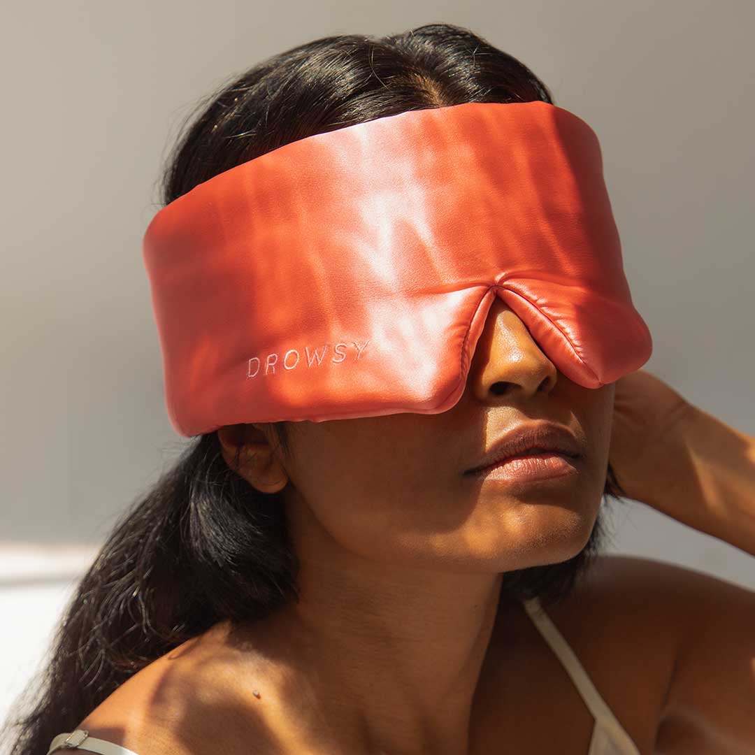 A woman wearing a shiny, padded red sleep mask labeled DROWSY sits in soft light, with her long dark hair visible and her face partially in shadow.