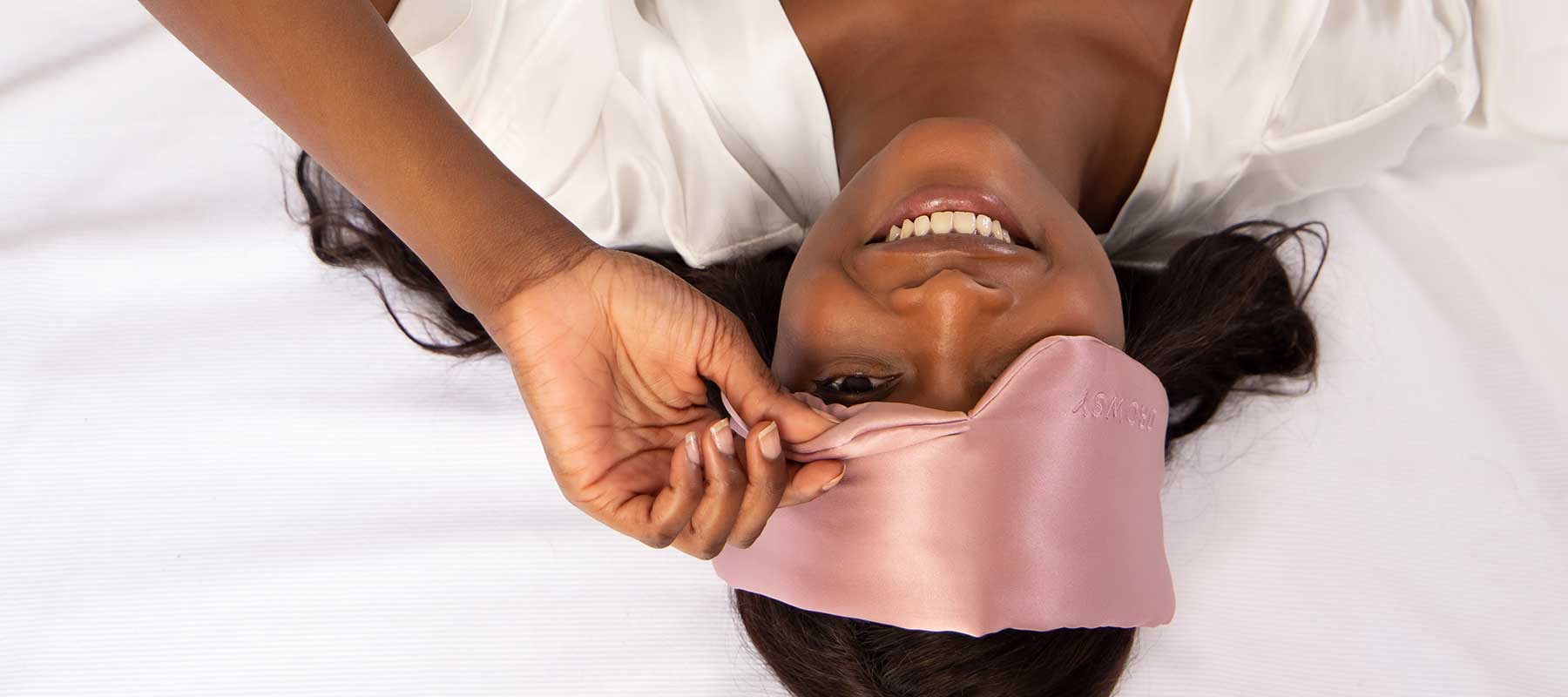 A woman lying on a bed, smiling, and lifting a pink satin sleep mask from her eyes while wearing a white robe.