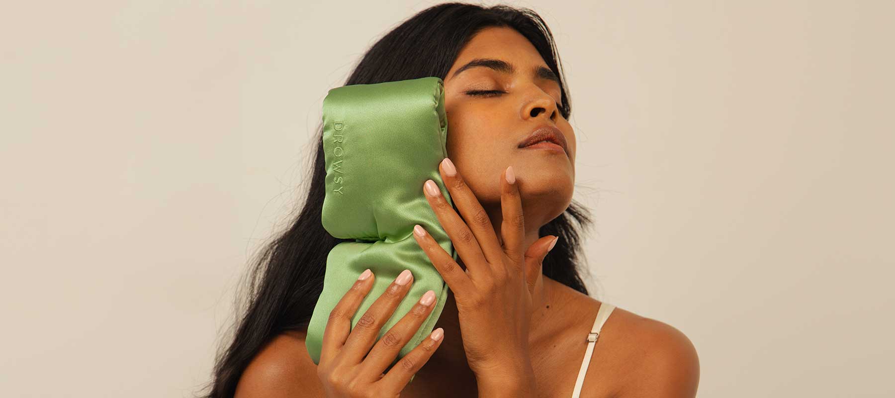 A woman with long dark hair presses a green satin ice pack to the side of her face, eyes closed, wearing a beige spaghetti strap top against a neutral background.