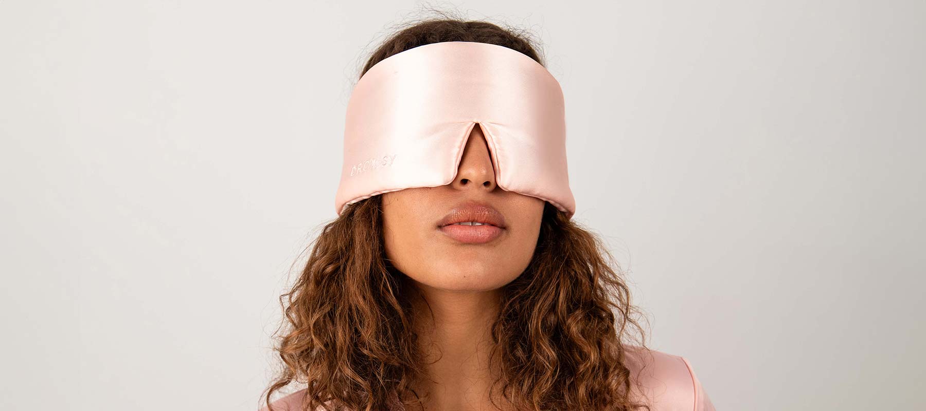 A person with curly brown hair wears a large, light pink silk sleep mask that covers their eyes and part of their nose, with a small slit in the center. The background is plain and light-colored.