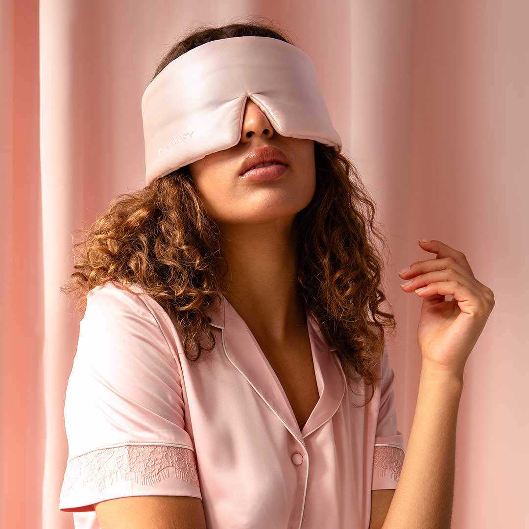 A woman with curly hair wears a pink satin sleep mask and matching pink satin pajamas, sitting against a pale pink background with her hand raised slightly near her face.