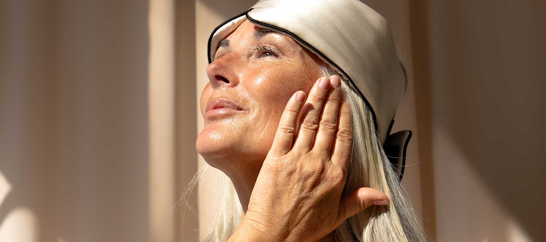 A woman with long gray hair and a satin sleep mask on her head touches her face while gazing upward, bathed in warm natural light.