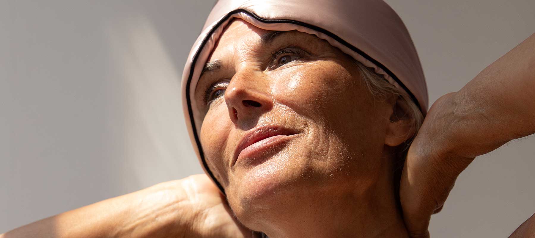 A close-up of an older woman wearing a light pink sleep mask pushed up on her forehead, looking up and smiling softly with her hands behind her neck, bathed in natural sunlight.