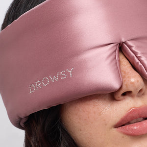 Close-up of a woman wearing the Damask Rose silk sleep mask by drowsysleepco, featuring a Swarovski®-embellished logo on the side. The soft pink mask comfortably covers her eyes and part of her nose, ensuring undisturbed beauty rest.