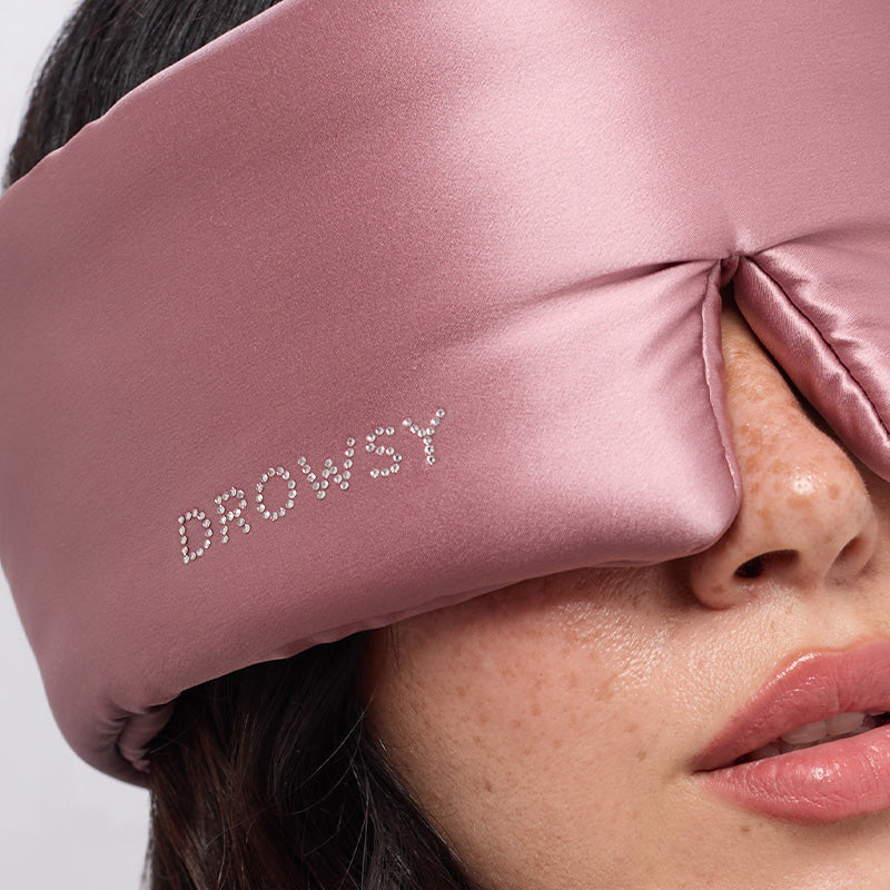 Close-up of a woman wearing the Damask Rose silk sleep mask by drowsysleepco, featuring a Swarovski®-embellished logo on the side. The soft pink mask comfortably covers her eyes and part of her nose, ensuring undisturbed beauty rest.